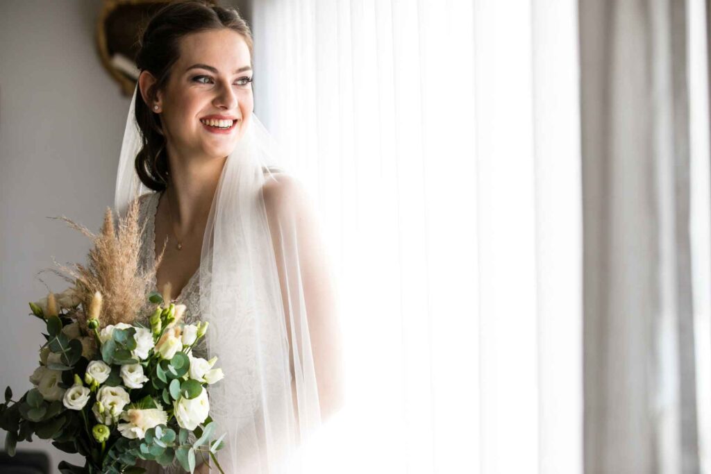 Die Braut in ihrem weißen Kleid steht entspannt vor einem großen Fenster und lässt sich von den warmen Sonnenstrahlen küssen, während sie sich auf ihren großen Tag vorbereitet. Der Raum ist mit Blumen und Accessoires dekoriert und die Vorfreude ist spürbar. Ein wunderschönes und emotionales Bild, das die Vorfreude und den Zauber des Hochzeitstages einfängt.